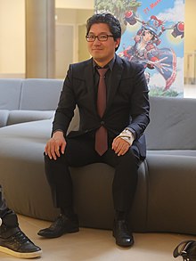 Photo de Yuji Naka, souriant et assis sur un canapé, en costume noir