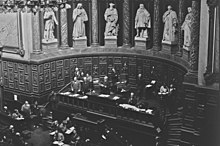 Photo en noir et blanc d'un hémicycle avec en hauteur un homme, cheveux blancs et barbe blanche, prononçant un discours