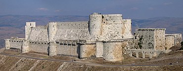 A stone castle with two high curtain walls, one within the other. They are crenelated and studded with projecting towers, both rectangular and rounded. The castle is on a promontory high above the surrounding landscape.