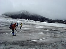 A black cone-shaped mountain rising over glacial ice and three climbers in the foreground.