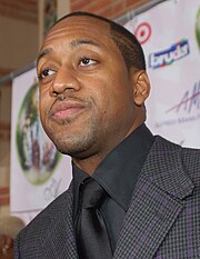 An African-American man with short hair and a stubble, wearing a black suit and tie.