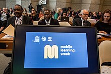 A computer screen at the front of a room of policy makers shows the Mobile Learning Week logo