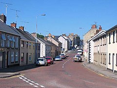 A road junction at the bottom of a hill, the adjourning road at the bottom to the left and the other road is climbing in the distance, surrounded by residential and commercial buildings.