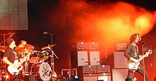 Photographie de deux guitaristes et un batteur lors d'un concert.