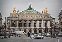 Frontale Farbfotografie eines zweigeschossigen Gebäudes im Neobarock-Stil. Im Erdgeschoss sind Rundbögen und im Obergeschoss sind zwischen den Fenstern Säulen, über denen „Académie nationale de musique“ steht. An den Dachecken stehen zwei goldene Figuren und das Gesims ist Gold. In der Mitte ist eine grüne Kuppel mit Figuren, die eine goldene Krone hochhalten.