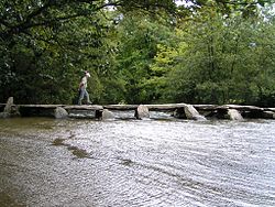 Low bridge of stone slabs supported by seven groups of vertical stones, across water with trees in the background.