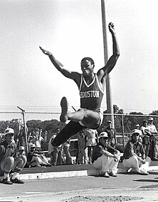 A man midway through a long jump leap. There is a metal chainlink fence in the background, both in front of which and behind are a number of spectators