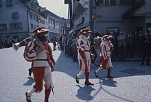 Photographie de trois hommes, portant la barbe et un costume en rouge et blanc, marchant dans une rue.