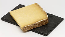 Une tranche carrée de cantal entre-deux, un fromage de couleur jaune à croûte marron. La croûte se trouve sur deux bords contiguës. Le fromage est présenté sur une ardoise le tout sur fond blanc.