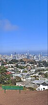 A view of downtown San Francisco from the top of a tall hill. In the foreground, a park bench on a dirt ground. In the background, a view of residential and commercial buildings in San Francisco, including skyscrapers further in the distance.