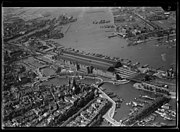 Luchtfoto van het IJ en omgeving, gezien in noordwestelijke richting. Op de voorgrond het het Oosterdok en het Stationseiland, op de achtergrond Amsterdam-Noord; circa 1930. Luchtvaartafdeeling, 1920-1940.