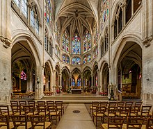 Frontale Farbfotografie eines inneren Kirchenschiffs mit einem schlichten Altar im Hintergrund. Vorne sind an beiden Seiten Holzstuhlreihen. Durch Spitzbögen, die den Raum komplett umrunden, sind beide Seitenschiffe mit kleinen Altären und Sitzen erreichbar. In der oberen Bildhälfte sind an allen Wandseiten Kirchenfenster mit farbigen Szenen und ein Deckengewölbe mit Spitzbögen.