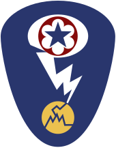 Badge d'épaule ovale avec un fond bleu foncé. Au sommet se trouve une étoile bleue entourée d'un cercle rouge qui correspond à l'écusson de l'Army Service Forces. Ce dernier est entouré d'un ovale blanc représentant un nuage en champignon. Un éclair blanc sort du nuage et frappe un cercle jaune fissuré représentant un atome.