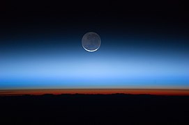 Månen i sentrum; jordens overflate nederst i overgang til den oransje-fargede troposfæren. Troposfæren har en skarp overgang til tropopausen, som vises som grensen mellom oransje- og blåfarget atmosfære.