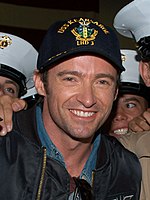 Um homem sorri para a câmera que ele usa uma jaqueta de cor escura sobre uma camisa jeans e um boné que possui um emblema azul-marinho. Ele está cercado por figuras vestidas de branco pico tampas, que são na sua maioria cortadas fora da vista.