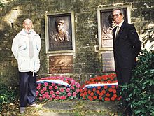 Photographie en couleur montrant deux hommes d'âge mûr se recueillant devant une stèle et des gerbes de fleur.