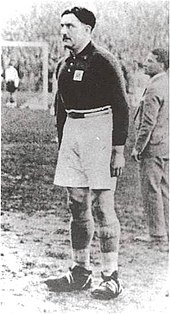 Photographie en noir et blanc d'un joueur de football debout sur un terrain, les bras le long du corps.