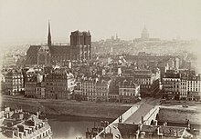 Schwarzweißfotografie in der Obersicht von einer Insel mit vielen alten Gebäuden und einer Brücke im Vordergrund, die über den Fluss führt. Links zwischen den Häusern steht eine Kathedrale mit zwei Ecktürmen und einem gotischen Turm auf dem Kirchenschiff. Im rechten Hintergrund, der nebelig erscheint, ist ein Kuppelbau.