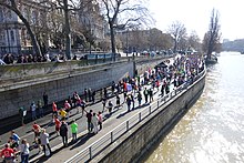 Seitliche Farbfotografie einer Straße, die links vom Ufer liegt und auf der viele Menschen von oben rechts nach unten links rennen. Auf den Gehwegen und auf einer erhöhten Mauer oben links stehen Zuschauer. Im Hintergrund sind Häuser und Bäume.