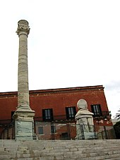 au-dessus d'un large escalier sont disposées une colonne entière et une base de colonne ; au fond, un bâtiment rouge.