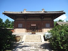 Fotografie budovy s typickou čínskou střechou se zvýšenými rohy, stavba stojí na zvýšené terase k níž vede schodiště, uprostřed vchodové dveře, vlevo a vpravo velké okno, v pozadí a po stranách v popředí jsou zelené stromy