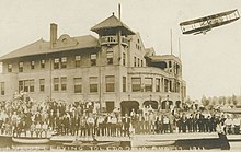 A postcard of Harry Atwood flying away from Toledo Ohio. The image depicts Atwood already in his plane in flight, while a crowd in front of a building faces the photographer.
