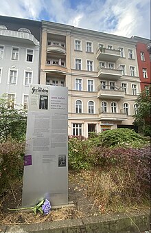 Im Vordergrund steht eine rechteckige, silberfarbene Tafel, die ein Foto von Lotte Hahm in der Zeitschrift „Die Freundin“ zeigt. Im Hintergrund sind Häuserfronten und grüne Vegetation zu sehen.
