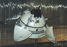 Zdjęcie. Model w muzeum. Okrągła kapsuła z rozłożonymi pokrywami i wystającymi antenami.