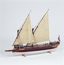 A side view of a model of a small galley with two masts rigged with lateen (triangular) sails. Its outrigger folded up and the oars stowed on the deck. The hull above the waterline is painted red with decorative details in gold and blue. The bow has a raised platform (rambade) armed with 3 small cannons.