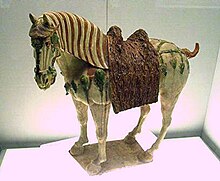 Fotografie keramické figurky osedlaného koně s trojbarevnou glazurou, kůň je světle béžový, hříva s hnědými pruhy, sedlo a sedlové pokrývky hnědé, ozdobné řemení zelenomodré