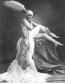 Photographie en noir et blanc d'une danseuse de music-hall en position assise, vêtue d'une robe longue et coiffée d'un chapeau à plumes.