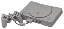 Photo d'une console grise, dotée d'un couvercle pour CD, de deux boutons à gauche et d'un bouton à droite. Une manette est branchée.