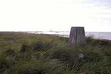 A small concrete pillar sits amongst long grass. There is a body of water in the background.