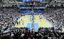 Packed basketball Arena, shown from behind one basket, looking across the court