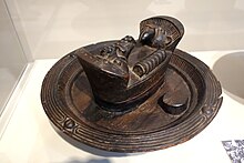 An image of a kola nut bowl in a museum showcase, it is wooden, round and brown and a small compartment is at the centre with a lid featuring animal faces carved on.