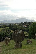Photographie d’un cimetière, avec des stèles discoïdales, sur fond de montagnes.