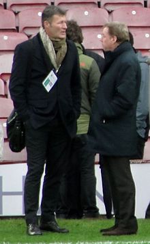 Two men wearing mostly dark coloured clothing stand at the side of a football pitch, hands in pockets, staring past each other.
