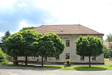 Patrová budova. V jejím čele (částečně zakryt košatými listnatými stromy, které před budovou rostou) je nápis „Obecná škola“.