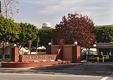 Entrée d'un campus avec un muret en brique rouge orné du nom Walt Disney Studios, derrière se profilent des arbres et des bâtiments colorés de plusieurs étages.