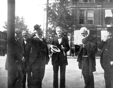 Fotografia in bianco e nero di sei uomini in piedi: sono tutti ben vestiti in giacca e cravatta. Cope ha i capelli corti, i baffi e una piccola barba; tra le mani tiene un cappello a tesa larga e dei fogli.