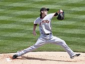 A man in a gray baseball uniform and black baseball cap throws a baseball with his right hand from a dirt mound on a grass field.