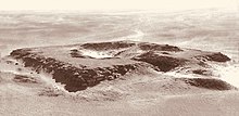 Black and white aerial photography of the ruins of a square-shaped monument with a depression in the center.