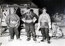 Photographie noir et blanc de trois hommes en tenue pour la marche (raquette, manteau, bonnet).