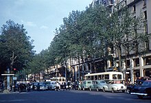 Seitliche Farbfotografie von einer viel befahrenen Allee mit Fußgängern, die eine linke Ampel überqueren. Rechts stehen vor Häuserfassaden zwei Omnibusse.