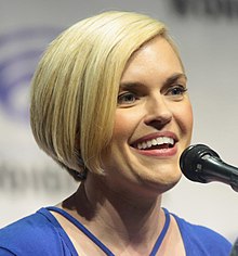 A blonde-haired young woman speaking into a microphone with a blue dress on.