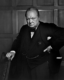 Churchill, 67, wearing a suit, standing and holding into the back of a chair