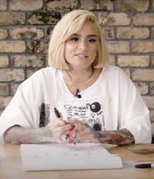 Kehlani smiles lightly, holding a permanent marker and drawing on a large pad of paper