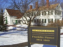 Photographie d'une maison blanche à deux étages avec de hautes cheminées en brique. Le sol est recouvert de neige et un panneau indique qu'il s'agit de la résidence du 14e président des États-Unis.