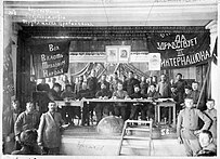 Photographie en noir et blanc d'une réunion, avec au centre un bureau sur lequel sont assis derrières de nombreuses personnes. Les murs ont des inscriptions en russes.