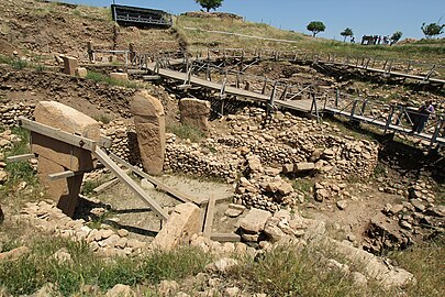 Vue sur un enclos circulaire entouré de murets de pierres. À l'arrière plan une passerelle est construite au-dessus d'une autre partie du chantier de fouilles.